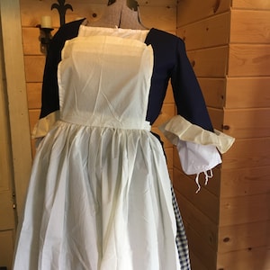 May include: A historical costume displayed on a mannequin. The outfit includes a navy blue bodice with long sleeves, a white apron, and a full white skirt. The sleeves have ruffled cuffs. The costume is reminiscent of colonial era clothing.
