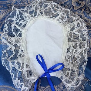 Puede incluir: Collar de encaje blanco con una cinta de satén azul atada en un lazo. El collar está sobre un fondo de tela con estampado azul y blanco.