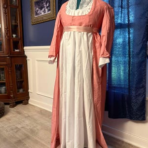 May include: A pink and white dress with a lace collar and a pink sash. The dress is long and flowing, and it has a simple, elegant design.