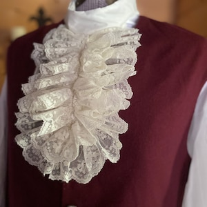May include: A white lace cravat with a ruffled design. The cravat is worn over a white shirt and a burgundy vest.