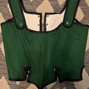 May include: A green corset with black trim and white lining. The corset has adjustable straps and a hook and eye closure in the front.
