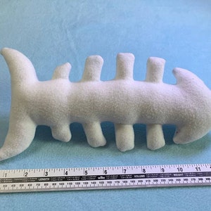May include: White plush toy shaped like a fish with a segmented body. The toy is approximately 10 inches long.