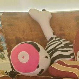 May include: A selection of plush food-shaped cushions. The collection features a white and black zebra-striped drumstick, a round pillow with a pink centre, and a cushion resembling bacon with red and gold stripes.