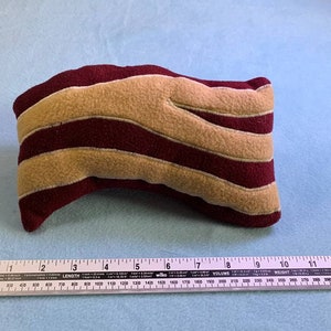 Puede incluir: Un juguete de peluche con forma de una rebanada de tocino. El juguete está hecho de tela roja y beige y mide aproximadamente 20 cm de largo.