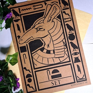Op de afbeelding: Een bruine papieren print met een zwart-witte illustratie van de Egyptische god Seth. De illustratie toont Seth als een jakhals met een mensachtig hoofd en een kroon. De print is omringd door hiërogliefen en het woord "SET" staat onder de illustratie. De handtekening van de kunstenaar "RYNOZERUS '18" staat in de rechterbenedenhoek.