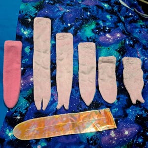 May include: Six pink fabric finger puppets of varying sizes, arranged in a row on a blue and purple galaxy print fabric. A long, iridescent pink and gold pouch is lying below the puppets.