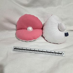 Op de afbeelding: Twee pluche schelpen van stof, een roze en een witte, met een parel in de roze schelp.