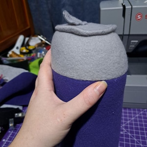 May include: A gray and purple plush toy shaped like a can. The top of the can is gray and the bottom is purple.