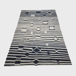 Pu&ograve; includere: Un tappeto rettangolare con sfondo color crema e motivi geometrici neri. Il design presenta strisce orizzontali intervallate da forme a diamante, quadrate e ovali. Il tappeto &egrave; esposto su una superficie bianca.