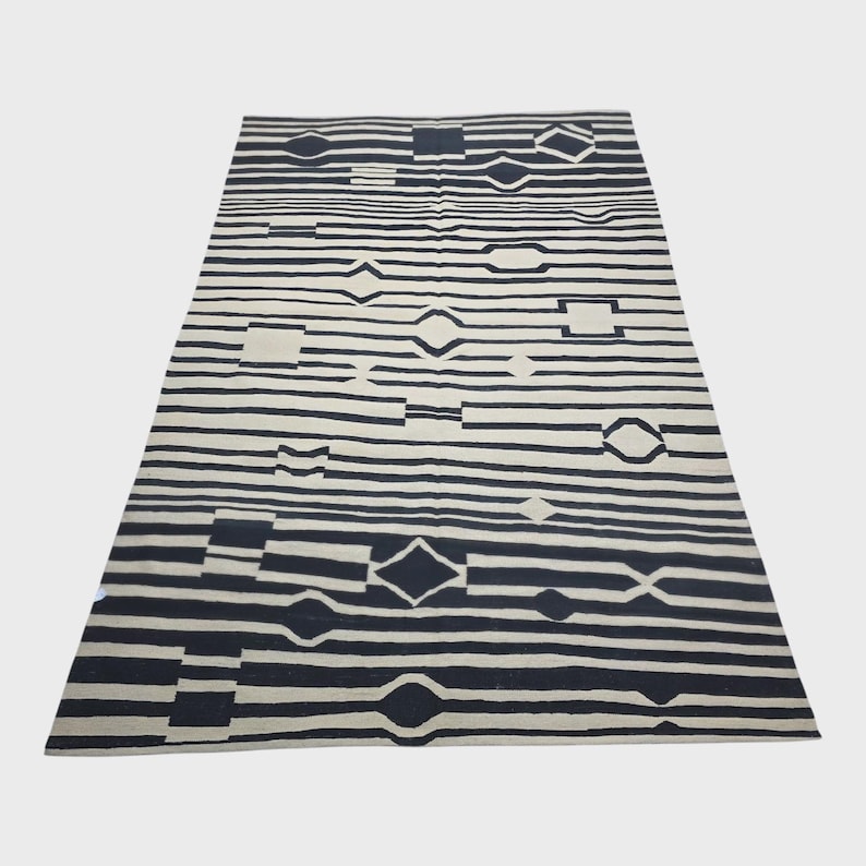 Pu&ograve; includere: Un tappeto rettangolare con sfondo color crema e motivi geometrici neri. Il design presenta strisce orizzontali intervallate da forme a diamante, quadrate e ovali. Il tappeto &egrave; esposto su una superficie bianca.