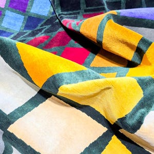 Puede incluir: Una alfombra colorida con patrones geom&eacute;tricos con una variedad de formas y colores. La alfombra presenta cuadrados, tri&aacute;ngulos y rombos en tonos de amarillo, morado, azul, rojo y gris. Los bordes est&aacute;n delineados en verde oscuro.