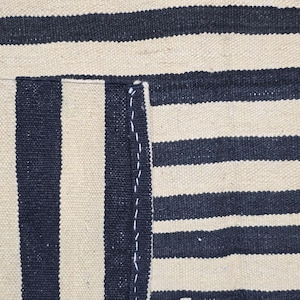 Pu&ograve; includere: Primo piano di un tessuto a trama fitta con strisce blu navy e beige alternate. Il tessuto ha un aspetto strutturato, con fili visibili e una cucitura sul lato sinistro. Le strisce sono distanziate uniformemente.