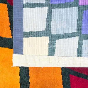 Puede incluir: Primer plano de una colorida alfombra geom&eacute;trica. La alfombra presenta un dise&ntilde;o de patchwork con cuadrados naranjas, rojos y morados. Un borde azul separa los cuadrados de la secci&oacute;n naranja. La alfombra tiene un aspecto texturizado y tejido.