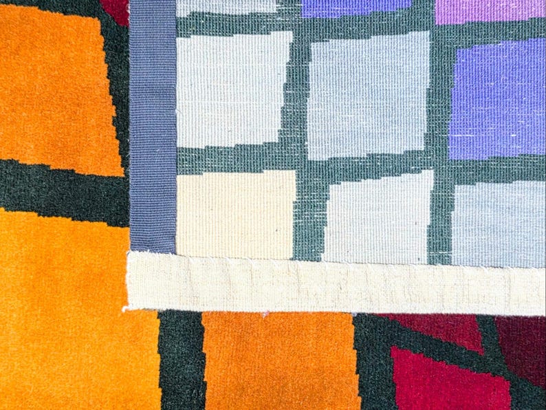 Puede incluir: Primer plano de una colorida alfombra geom&eacute;trica. La alfombra presenta un dise&ntilde;o de patchwork con cuadrados naranjas, rojos y morados. Un borde azul separa los cuadrados de la secci&oacute;n naranja. La alfombra tiene un aspecto texturizado y tejido.