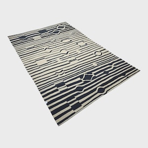 Pu&ograve; includere: Un tappeto rettangolare con un motivo geometrico. Il tappeto presenta strisce orizzontali alternate in bianco sporco e blu navy. Il design include forme a diamante, quadrate ed esagonali. Il tappeto &egrave; realizzato in materiale morbido.