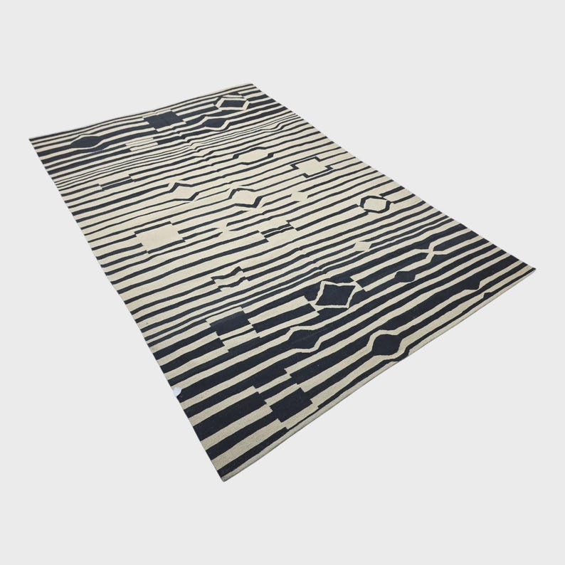 Pu&ograve; includere: Un tappeto rettangolare con un motivo geometrico. Il tappeto presenta strisce orizzontali alternate in bianco sporco e blu navy. Il design include forme a diamante, quadrate ed esagonali. Il tappeto &egrave; realizzato in materiale morbido.