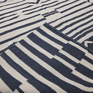 Pu&ograve; includere: Un tappeto con un motivo a righe blu navy e bianco sporco. Le strisce sono orizzontali e di larghezze variabili, creando un design geometrico. Il tappeto &egrave; piegato, rivelando il lato inferiore.