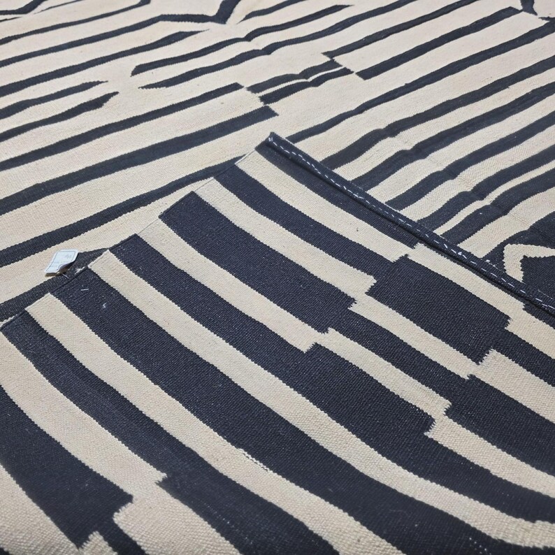 Pu&ograve; includere: Un tappeto con un motivo a righe blu navy e bianco sporco. Le strisce sono orizzontali e di larghezze variabili, creando un design geometrico. Il tappeto &egrave; piegato, rivelando il lato inferiore.