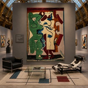 May include: A large wall tapestry featuring abstract green, white, and red figures, displayed above a beige rug with black lines and coloured squares. A black leather armchair and a cowhide chaise lounge sit on the rug with a glass coffee table.