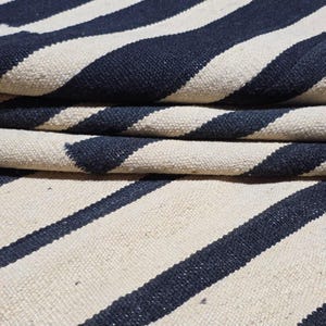 Pu&ograve; includere: Tappeto piegato con strisce blu navy e bianco sporco alternate. Il tappeto &egrave; realizzato in un materiale strutturato, con le strisce diagonali. Il tappeto &egrave; piegato, mostrando pi&ugrave; strati del motivo a strisce.
