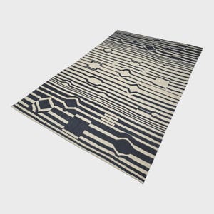 Pu&ograve; includere: Un tappeto rettangolare con un design geometrico. Presenta strisce alternate bianco sporco e blu navy. Il design include varie forme, come rettangoli e diamanti, creando un'estetica moderna e accattivante.