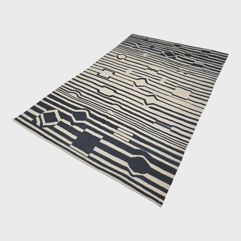 Pu&ograve; includere: Un tappeto rettangolare con un design geometrico. Presenta strisce alternate bianco sporco e blu navy. Il design include varie forme, come rettangoli e diamanti, creando un'estetica moderna e accattivante.
