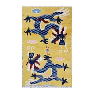 Op de afbeelding: Een rechthoekig geel tapijt met een herhaald patroon van twee blauwe draken met rode accenten. De draken zijn omgeven door blauwe en roze wolkvormige vormen. Het tapijt heeft een franje langs de onderkant.