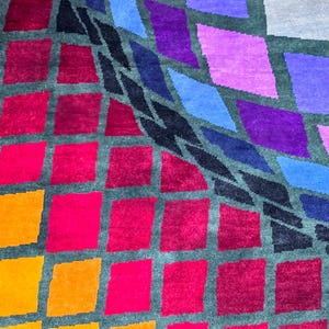 Puede incluir: Una colorida alfombra con un dise&ntilde;o geom&eacute;trico. El dise&ntilde;o presenta una cuadr&iacute;cula de formas de rombos en tonos de rojo, naranja, rosa, morado, azul y negro sobre un fondo gris. La alfombra tiene una apariencia texturizada.