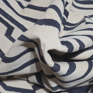 Pu&ograve; includere: Primo piano di un tappeto tessuto con sfondo color crema e strisce curve e a zig-zag blu navy. Il tappeto ha una superficie strutturata ed &egrave; piegato, mostrando la profondit&agrave; e il design del motivo.