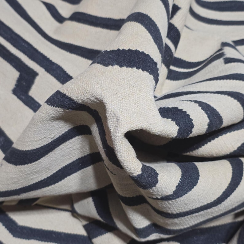 Pu&ograve; includere: Primo piano di un tappeto tessuto con sfondo color crema e strisce curve e a zig-zag blu navy. Il tappeto ha una superficie strutturata ed &egrave; piegato, mostrando la profondit&agrave; e il design del motivo.