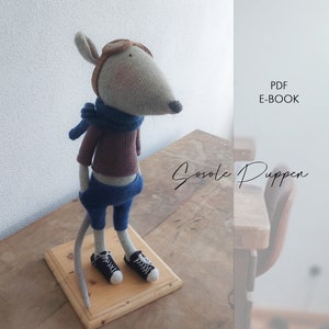 Könnte beinhalten: Eine gehäkelte graue Maus, die einen braunen Pullover, eine blaue Hose und einen blauen Schal trägt. Die Maus trägt eine Fliegerbrille und schwarze Turnschuhe. Die Maus steht auf einem Holzsockel. Der Text "PDF E-BOOK" und "Sosole Puppen" ist im Hintergrund sichtbar.