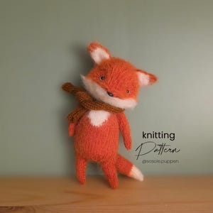 May include: A knitted orange fox toy with a white chest and ear tips, wearing a brown scarf. The fox has a small black nose and is posed against a muted green background. Text on the image reads "knitting Pattern @sosole.puppen".
