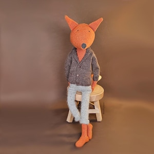 May include: A crocheted fox wearing a brown cardigan and gray pants sits on a wooden stool. The fox has orange fur and a black nose.