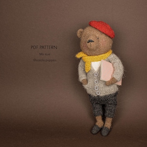 Puede incluir: Un muñeco oso tejido de color marrón con una boina roja, una bufanda amarilla, un cárdigan gris claro con botones y pantalones gris oscuro. El oso sostiene un pequeño libro a rayas rosas y blancas. Texto: PDF PATTERN MR BEAR @sosole.puppen.