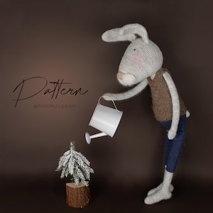 以下が含まれることがあります： 茶色のベストと青いズボンを着た、編み物の灰色のウサギの人形が、白いじょうろで木の台座にある小さな常緑樹に水をやっています。「Pattern」という単語とユーザー名「@sosole.puppen」が画像に書かれています。