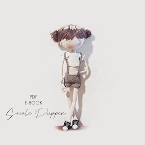 May include: Crochet doll with brown hair, a white crocheted top, brown shorts, and black shoes. The doll is wearing a flower crown and is standing against a white wall. The text "PDF E-BOOK Gosole Puppen" is visible in the image.