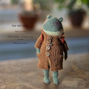 May include: A knitted green frog doll wearing a brown textured coat with four wooden buttons and a belt buckle. The frog has a small backpack and the text PDF PATTERN FROG TO KNIT COAT, BACKPACK, TROUSERS, SCARF TO KNIT @sosole.puppen is visible.