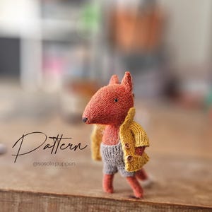 Könnte beinhalten: Eine gestrickte Fuchspuppe mit orangefarbenem Fell, die eine gelbe Jacke und graue Shorts trägt. Das Wort "Pattern" ist in Schreibschrift geschrieben. Die Puppe steht auf einer Holzoberfläche.