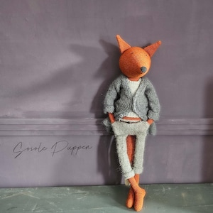 May include: A handmade fox doll with orange head and ears, grey sweater, trousers, and orange feet. The doll is leaning against a purple wall. The doll has a white shirt underneath the grey sweater. The doll is wearing a belt.