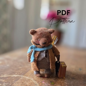 Könnte beinhalten: Ein handgefertigter brauner Teddybär mit blauem Schal und braunem Mantel, der einen kleinen Koffer und einen Regenschirm trägt. Der Bär hat ein Strick-Aussehen und der Text "PDF Pattern" ist sichtbar.