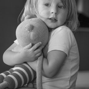 Puede incluir: Una niña pequeña con cabello rubio sostiene un animal de peluche de crochet a rayas. Lleva una camiseta blanca y tiene una expresión seria en su rostro.