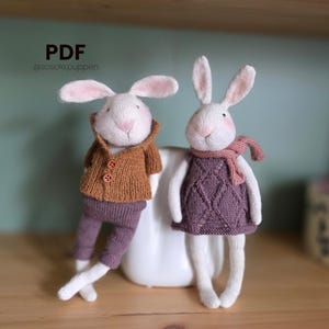 Op de afbeelding: Twee handgemaakte witte konijnenpoppen met roze oren en roze wangen. Eén konijn draagt een bruine trui en een paarse broek, de ander een paarse jurk en een roze sjaal. De afbeelding bevat de tekst "PDF @sosole.puppen".