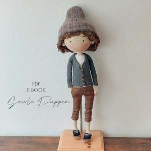 May include: Crochet doll pattern PDF e-book. The doll is wearing a brown hat, a green cardigan, brown pants, and black shoes. The doll is standing on a wooden base. The text "Gosolic Puppen" is written in black on a white background.