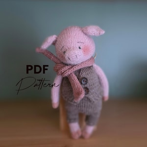 Puede incluir: Un cerdito rosa tejido a mano con un chaleco gris, pantalones y una bufanda rosa. El cerdito tiene mejillas rosadas y pequeños ojos negros. La imagen incluye el texto "PDF Pattern".