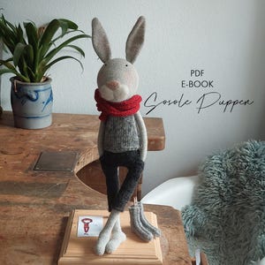 May include: A handmade stuffed rabbit doll with long ears, wearing a red scarf, gray sweater, and black pants. The doll is standing on a wooden base. The text "PDF E-BOOK Sosole Puppen" is visible.