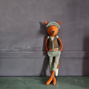 May include: A knitted fox doll wearing a brown waistcoat, grey trousers, and a blue hat. The doll is standing against a grey wall with the text "Sovole Puppen" written on it.