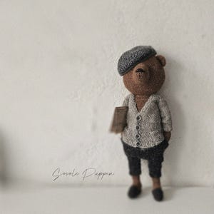 Puede incluir: Un oso de crochet marrón que lleva una boina gris y un cárdigan gris con botones. El oso sostiene una bolsa de papel marrón y lleva pantalones negros y zapatos negros. El oso está de pie sobre un fondo blanco. El texto "Sovole Puppen" está escrito en negro en la parte inferior de la imagen.