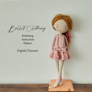 May include: A handcrafted crochet doll dressed in a pink ballet ensemble. The doll features auburn hair and wears a pink sweater, a fluffy skirt, and ballet shoes. The image includes the text "Ballet Clothing" and "Anleitung Instruction Pattern English/German".