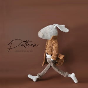 Könnte beinhalten: Ein gestricktes Spielzeugkaninchen, das einen braunen Mantel und eine Hose trägt. Das Kaninchen geht mit dem rechten Bein nach vorne. Der Hintergrund ist eine braune Wand. Der Text "Pattern @sosolepuppen" ist im Bild sichtbar.