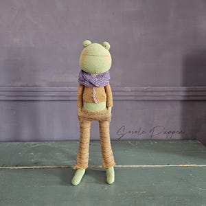 Könnte beinhalten: Eine grüne gehäkelte Froschpuppe, die einen lila Schal und eine braune und lila gestreifte Hose trägt. Die Puppe steht auf einer grünen Holzoberfläche. Der Text "Gosole Puppen" ist im Hintergrund sichtbar.
